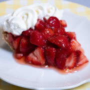 A slice of No Bake Strawberry Pie finished with whipped cream, sits on a white plate with a yellow gingham background.