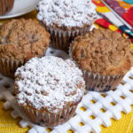 Four Blueberry Crumb Muffins sit on a white lattice plate with colored napkins and a yellow placemat.