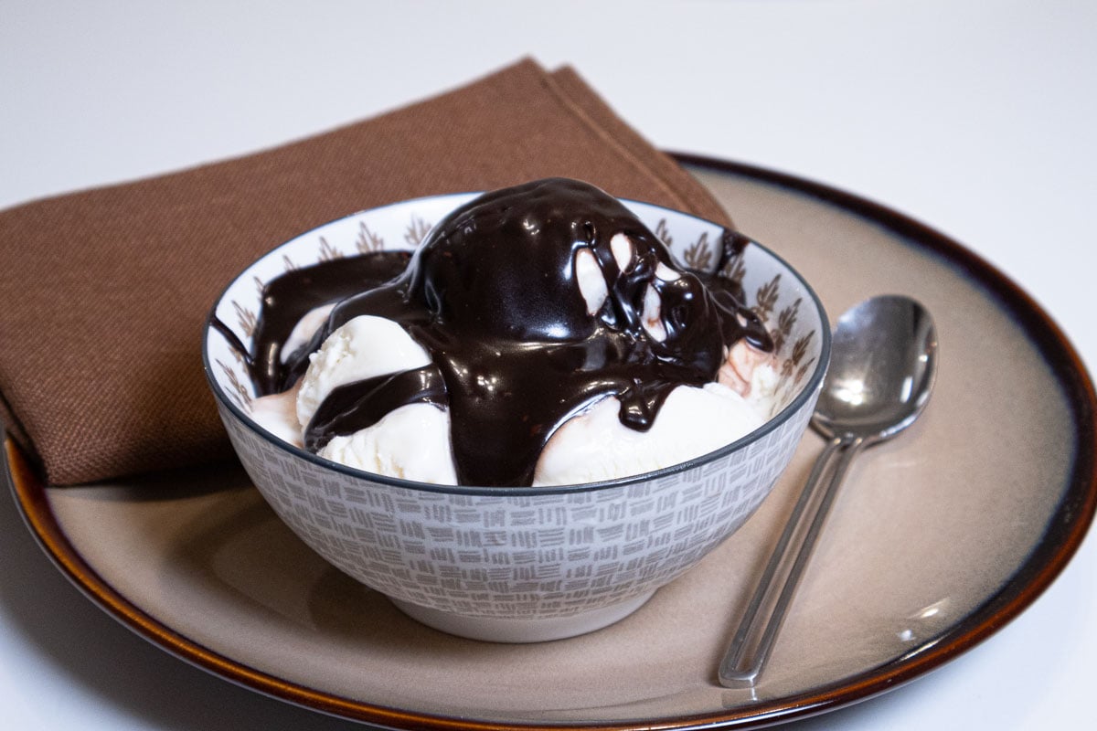 The bottom pictures shows the vanilla ice cream covered with the hot hot fudge sauce in a bowl on a beige plate with a brown napkin and spoon.