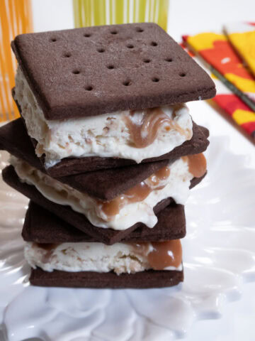 Three Ice Cream Sandwiches on a white plate with multi colored glasses and napkins behind.