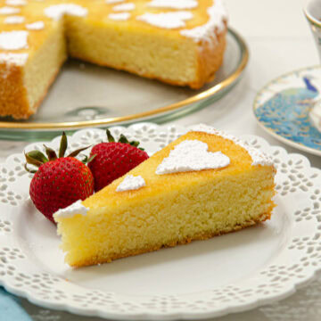 A slice of the Orange Almond Tea Cake sits on a white plate with 2 strawberries and the rest of the cut cake is behind it.