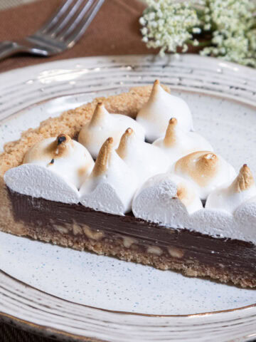 A slice of the S'more Tart with it's browned marshmallow topping sits on a brown rimmed plate with a fork and napkin in the background.