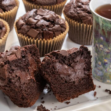 A cut open Double Chocolate Truffled Muffin sits on a plate with a teacup next to it and more muffins in the background.