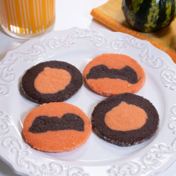 Four Halloween cookies on a white plate wih a glass of milk and several gourds in the background,