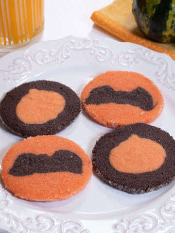 Four Halloween cookies on a white plate wih a glass of milk and several gourds in the background,