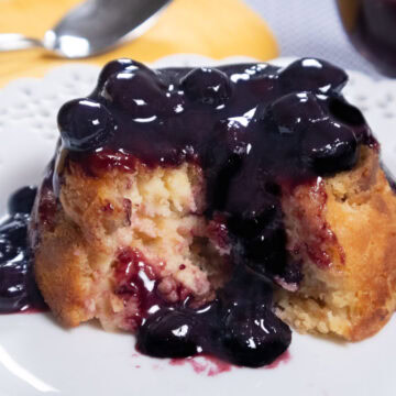 The Lemon Cheesecake Bread Pudding is shown partial opened up with a blubeberry sauce dripping down it on a white plate,