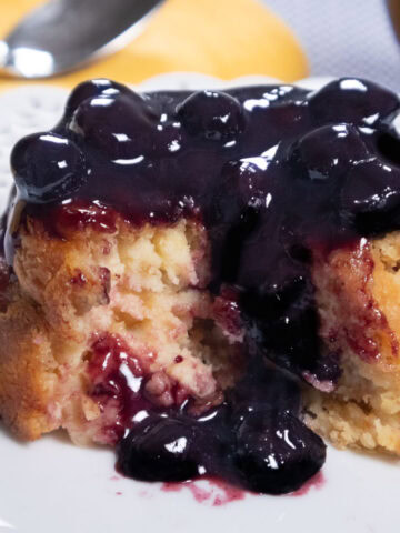 The Lemon Cheesecake Bread Pudding is shown partial opened up with a blubeberry sauce dripping down it on a white plate,