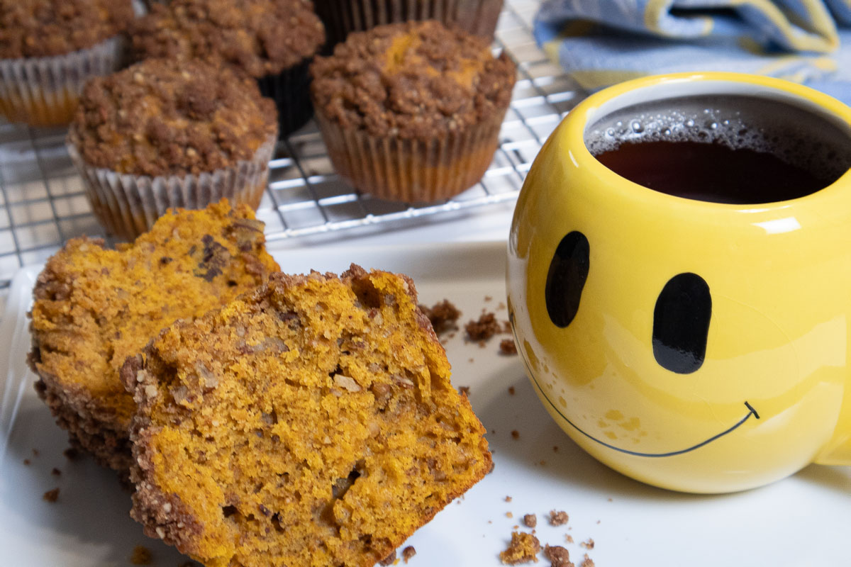 A broken muffin on a white plate with a yellow smile cup and muffins in the background