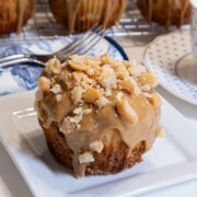 An Orange Macadamia Roll sits on a white plate with a rack of other rolls behind it and a saucer to a coffee cup to the right.