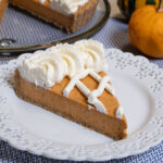 A slice of the pumpkin tart finished with a lattice design and whipped cream sits on a white plate with an orange and green gourd behind it as well as the pumpkin tart itself