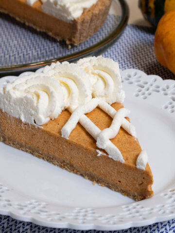 A slice of the pumpkin tart finished with a lattice design and whipped cream sits on a white plate with an orange and green gourd behind it as well as the pumpkin tart itself