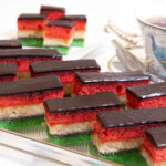 The featured picture shows a two rows on the diagonal of the Neapolitan cookies cut in bars some in the background cut in half and a cup of tea on the side.