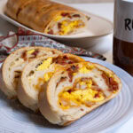 3 slices of Homemade Breakfast Stromboli sit on a wite plate with a mug and the remainder of the roll in the background.