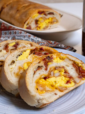 3 slices of Homemade Breakfast Stromboli sit on a wite plate with a mug and the remainder of the roll in the background.