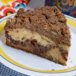 A slice of the Apple Cinnamon Coffee Cake sits on a yellow and blue rimmed plate with a multi-colored napkin and mug in the background.