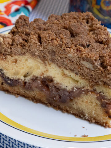 A slice of the Apple Cinnamon Coffee Cake sits on a yellow and blue rimmed plate with a multi-colored napkin and mug in the background.