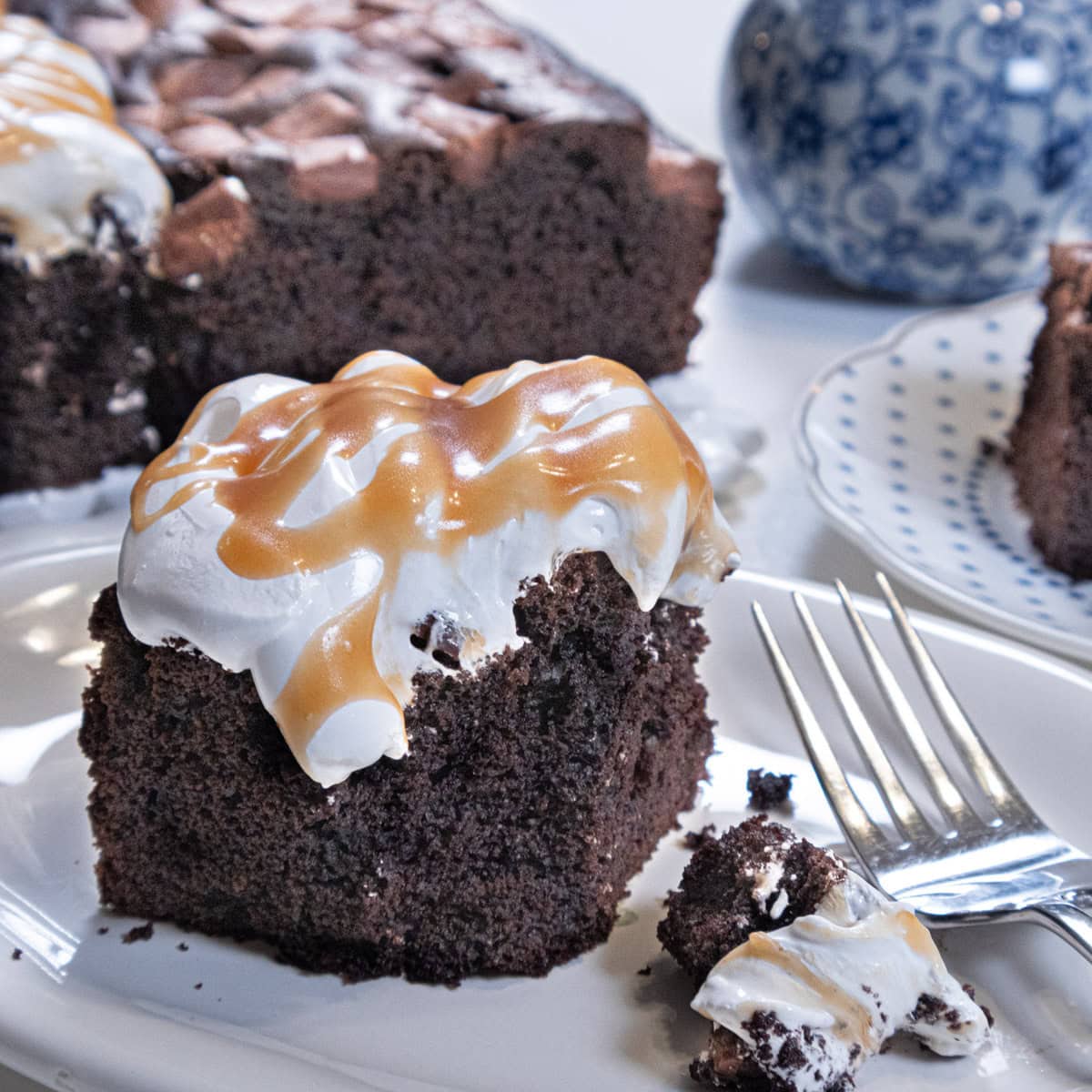 A partially eaten piece of the Decadent Chocolate Snack Cake sits on a white plate with a fork and bit of marshmallow with the cake in the background and a blue and white fase. 