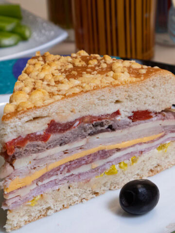 A slice Muffaletta Bread layered with meats, cheeses and condiments on a plate with olives and celery.