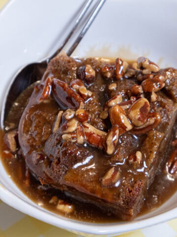 The Banana Bread Bread Pudding sits in a white bowl covered with the bourbon pecan sauce on a yellow and white background.