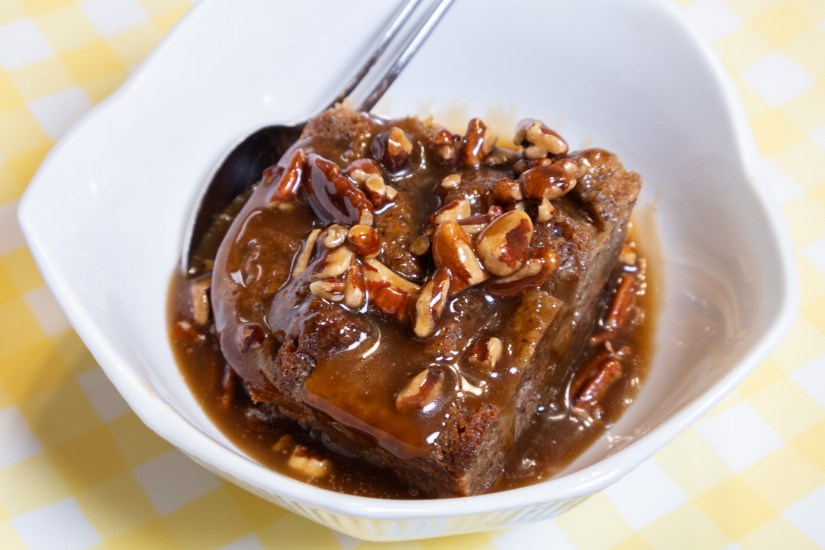 A portion of the Banana Bread Bread Pudding is in a white bowl with a spoon on a yellow and white background.