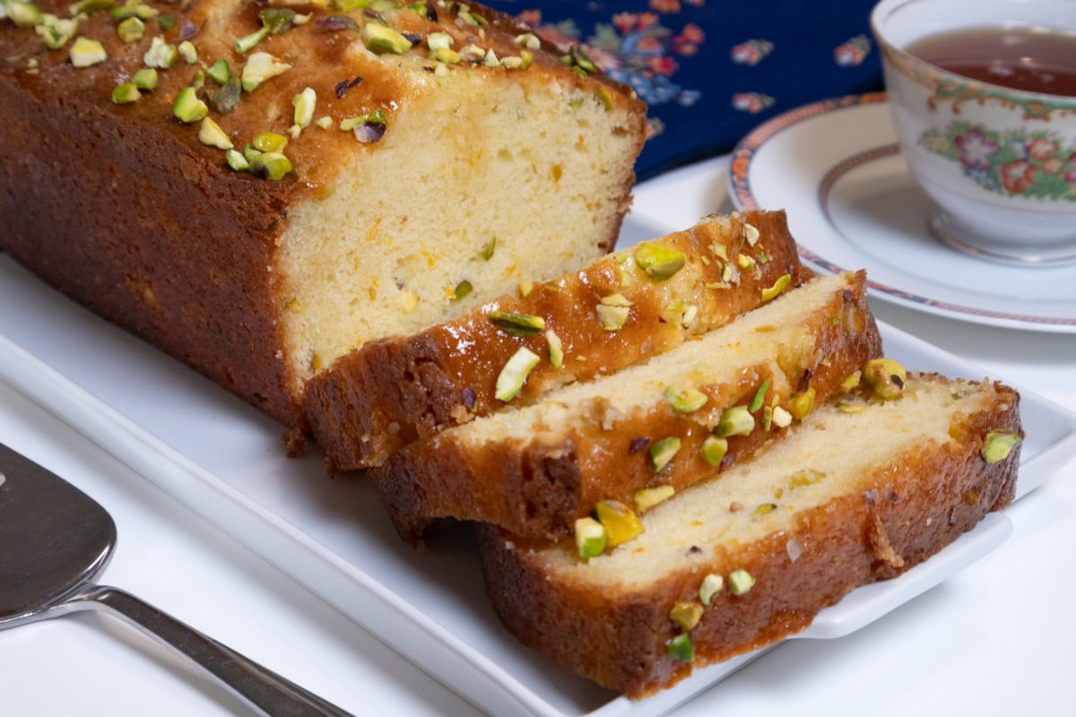 This photo of the Orange Pistachio Loaf Cake features 3 slices of cake on a white dish with the rest of the cake, blue printed napkins in the background with a cup of tea.
