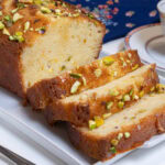 Slices of the Orange Pistachio Loaf Cake sit on a white tray with a blue napkin behind it and abcup of tea to the right.