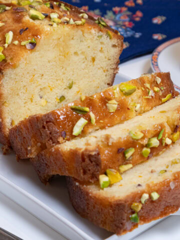 Slices of the Orange Pistachio Loaf Cake sit on a white tray with a blue napkin behind it and abcup of tea to the right.