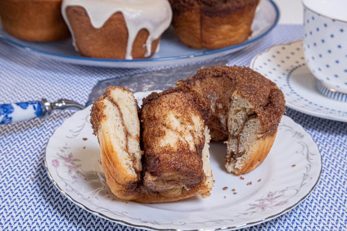 The open cinnamon crunch roll is on a white plate with flowers on the edge. A plate of rolls is in the background and a blue dotted cup and saucer are on the right. 