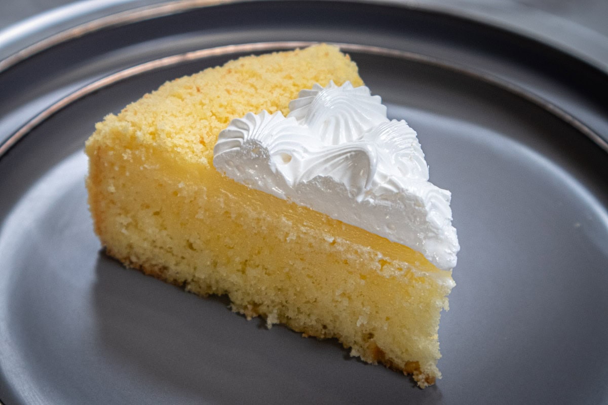 This picture shows a slice of the Triple Lemon Cake with the cake on the bottom, the lemon topping and the Swiss meringue finish sitting on a gray, gold rimmed plate.   