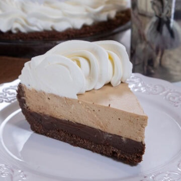 A slice of Bailey's Pie for Pi Day with its dark bottom layer, light mousse layer and whipped cream edging sits on a white plate with the ie in the background and a cup next to the plate.