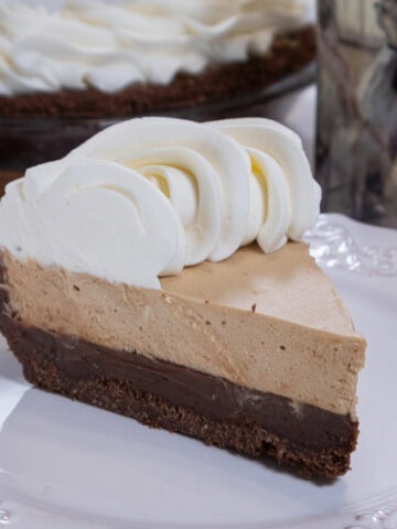 A slice of Bailey's Pie for Pi Day with its dark bottom layer, light mousse layer and whipped cream edging sits on a white plate with the ie in the background and a cup next to the plate.