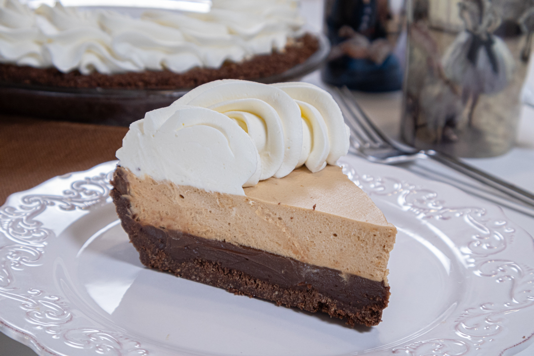 The photo shows a slice of the Bailey's pi day pie sits on a white plate with a whole pie in the back ground and two mugs and a fork.