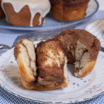 An open Cinnamon Roll sits on a flowral plate with a plate of rolls behind it and a blue dotted cup and saucer are off to the side.
