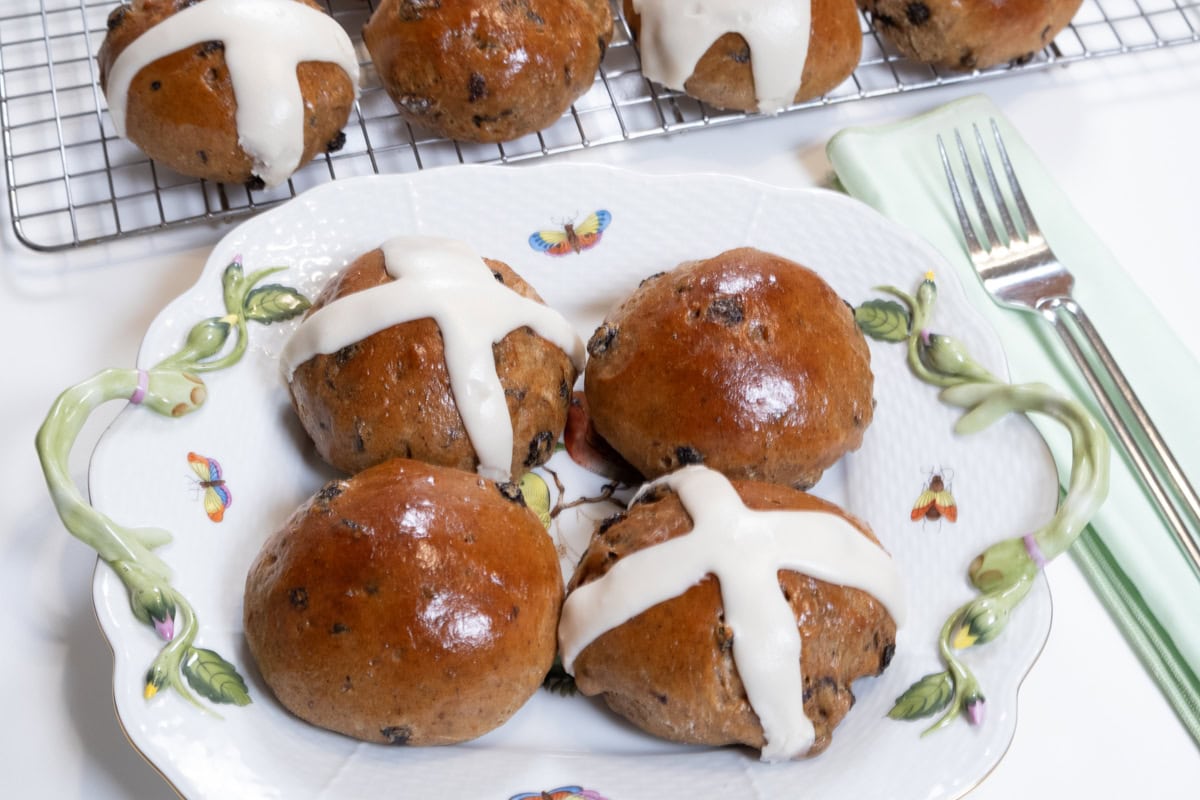 This last photograph of the Hot Cross Buns is on a white plate decorated with bees and flowers with more buns in the background on a cooling rack.