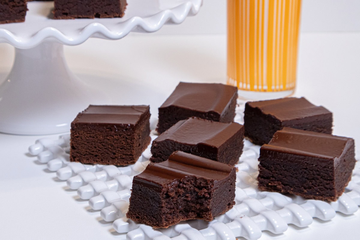 Several Mexican Chocolate brownies sit on a white lattice plate with a white cake plate with more brownies behind it and an orange glass of milk.
