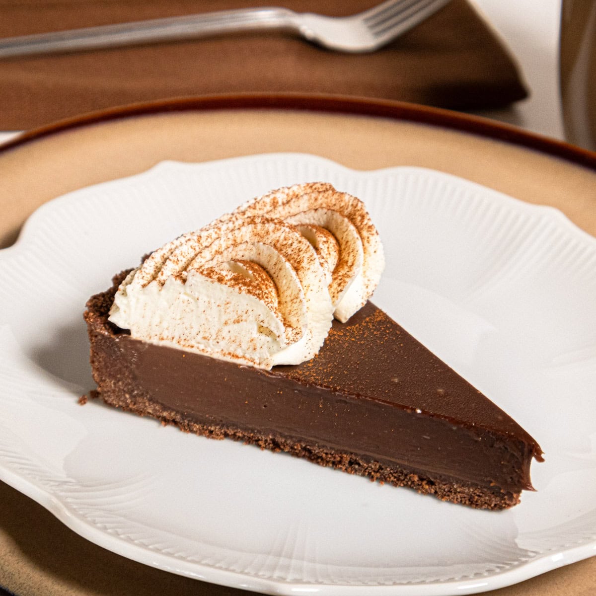 A slice of the Cafe au Lait tart sits on a white plate that is on a tan plate with a napkin and fork behind it.