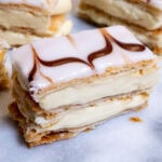 A slice of Mille-Feuille (Napoleon Pastry) with its white fondant and chocolate chevron desigh sits on a marble late with other slices in the background.