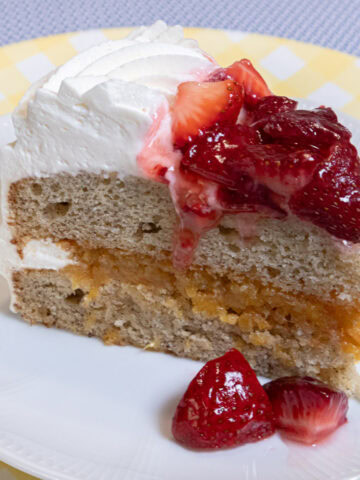 A slice of My Best Banana Cake Recipe sits on a white plate which is on a yellow gingham plate on a blue background.
