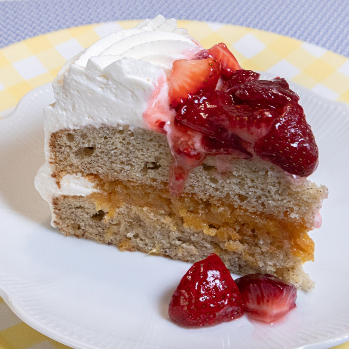 A slice of My Best Banana Cake Recipe sits on a white plate which is on a yellow gingham plate on a blue background.