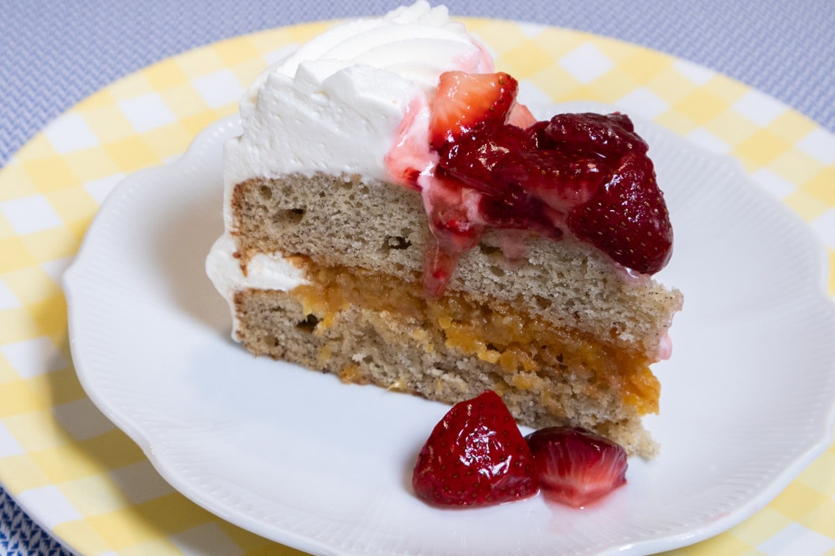 A slice of My Best Banana Cake Recipe sits on a white plate which is on a yellow gingham plate on a blue background.