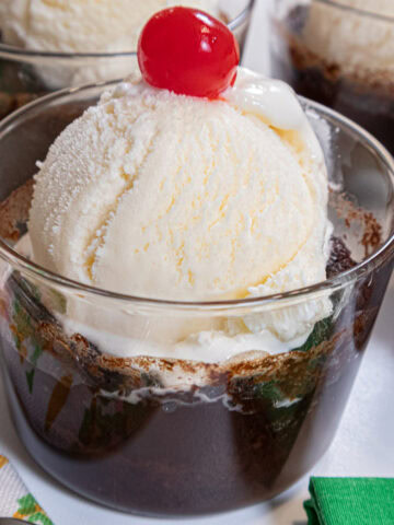 This Caramel Hot Fudge Sundae sits in a clear glass with a container with a scoop of vanilla ice cream with a cherry on top.