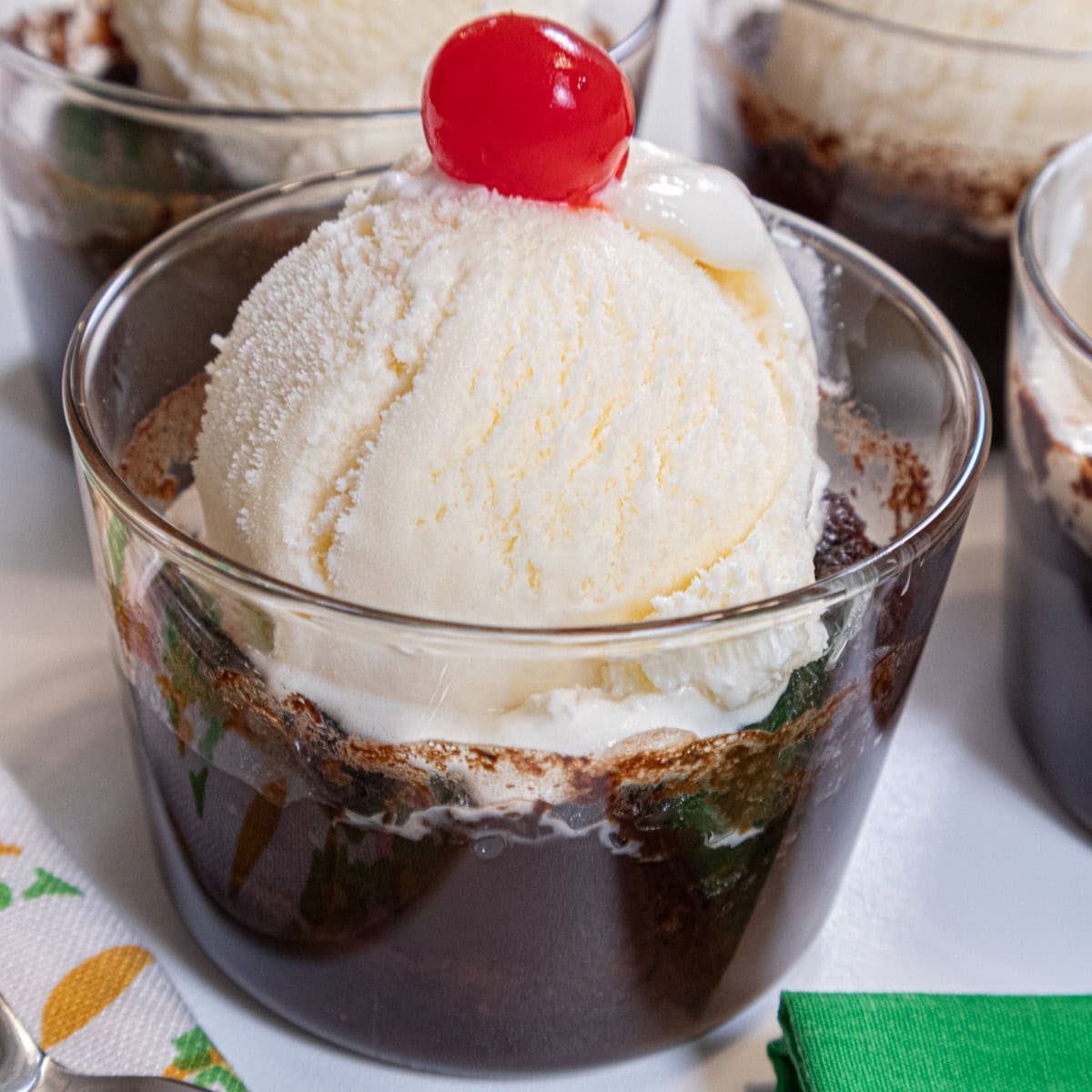 This Caramel Hot Fudge Sundae sits in a clear glass with a container with a scoop of vanilla ice cream with a cherry on top.