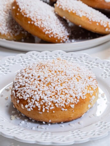 A single Lemon Sugar Bun sits on a white laced edged plate with a platter of more buns in the background.