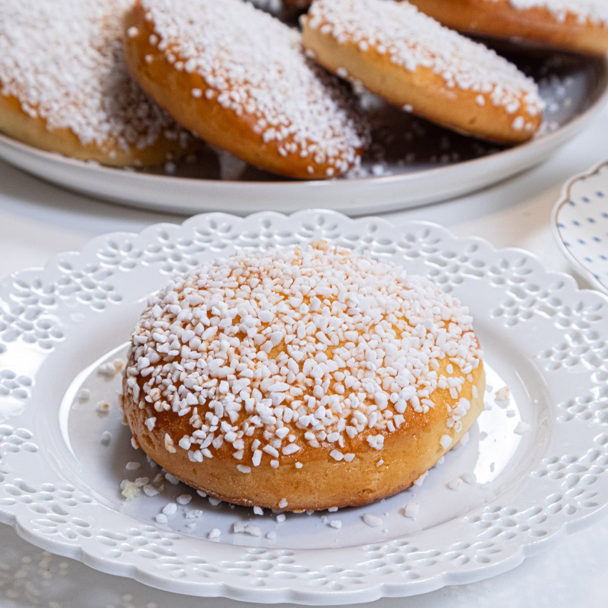 A single Lemon Sugar Bun sits on a white laced edged plate with a platter of more buns in the background.