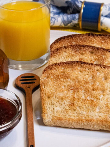 Slices of English Muffin Bread on a white plate with orange juice, honey and jam with a blue and white napkin in the background.