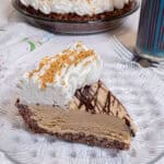 A slice of the Peanut Butter Ice Cream Pie sits on a clear plate over a lattice trivet with the pie in the background and a glass of ice tea nearby .