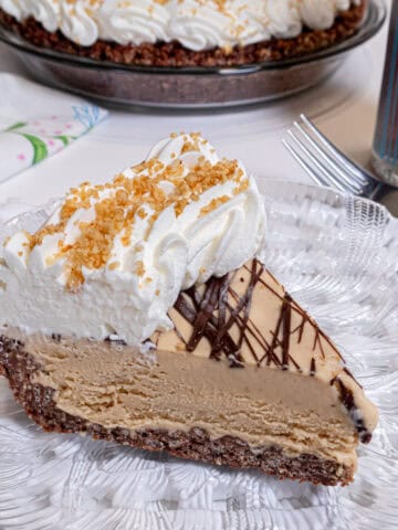 A slice of the Peanut Butter Ice Cream Pie sits on a clear plate over a lattice trivet with the pie in the background and a glass of ice tea nearby .