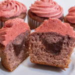 An open Strawberry Surprise Cupcake is cut open to display the chocolate inside with more cupcakes in the background.