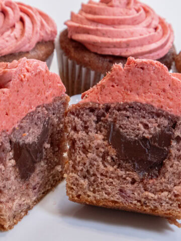 An open Strawberry Surprise Cupcake is cut open to display the chocolate inside with more cupcakes in the background.