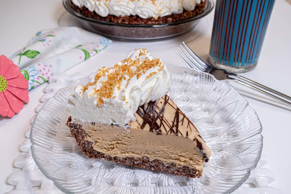 A slice of the Peanut Butter Ice Cream Pie sits on a clear plate over a lattice trivet with the pie in the background and a glass of ice tea nearby .